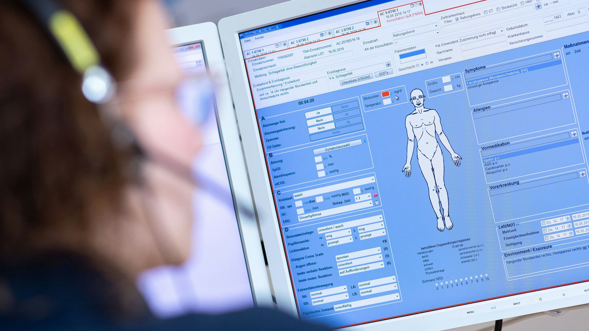 Eine Telenotärztin sitzt in der Rettungsleitstelle der Feuerwehr vor einem Bildschirm und ist mit Rettungssanitätern verbunden, die eine Schlaganfallpatientin behandeln.