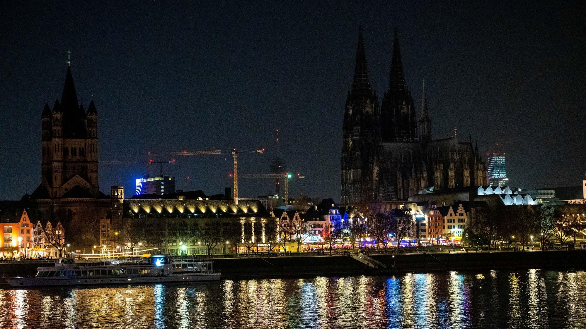 Das Bild zeigt die dunkle Skyline Kölns mit wenigen beleuchteten Altstadthäusern.