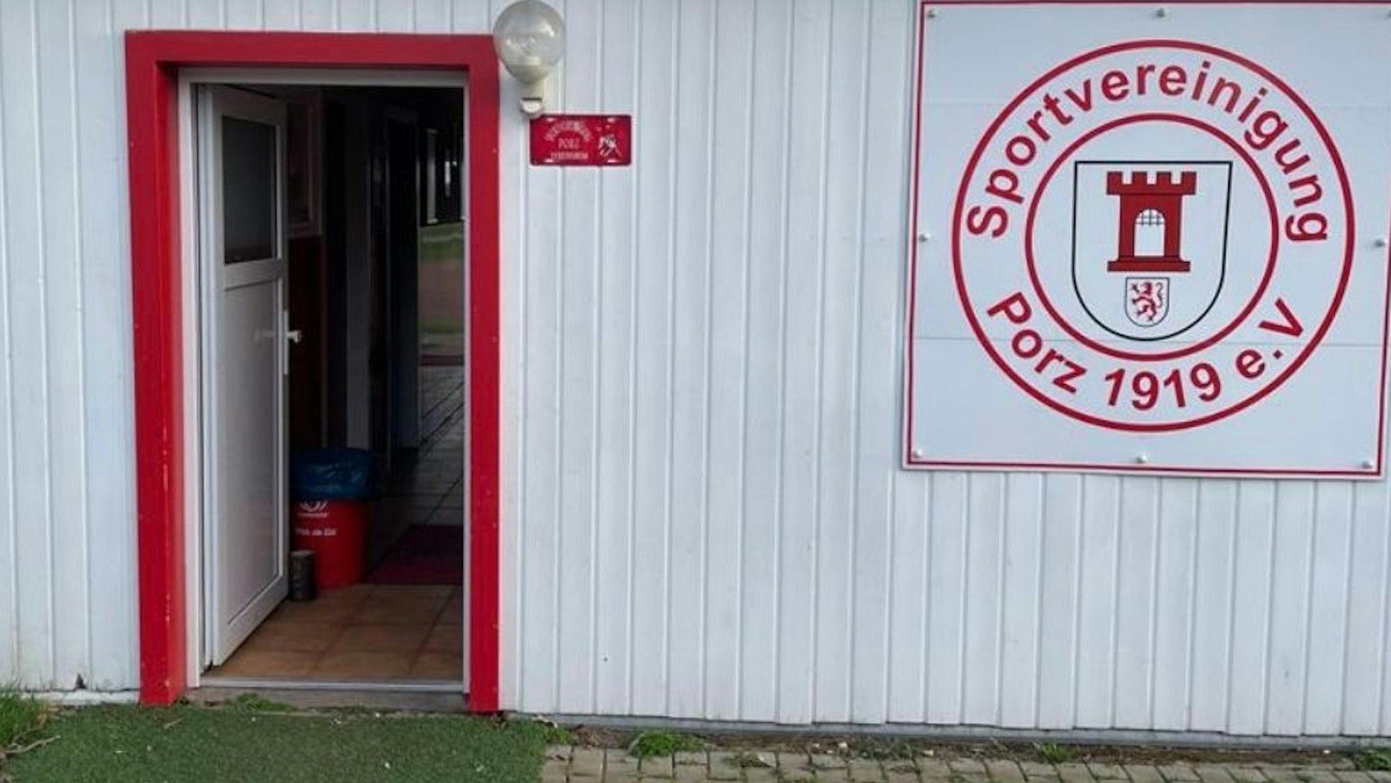 Eingang vom Vereinsheim der Sportvereinigung Porz an der Brucknerstraße. Weiße Fassade mit roten Akzenten und Logo.