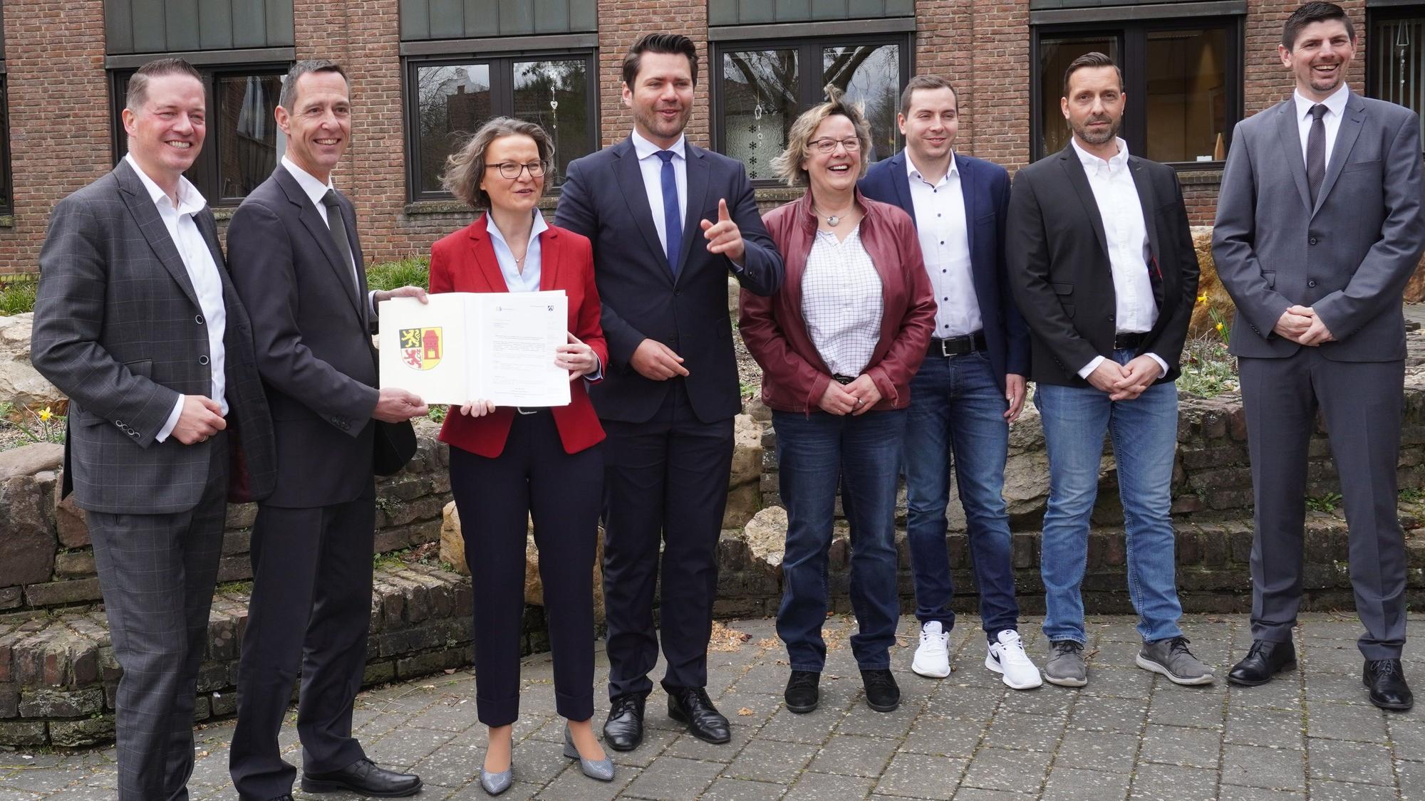 Eine Gruppe Männer und Frauen lächelt in die Kamera. Die NRW-Ministerin Ina Scharrenbach hält einen Förderbescheid.