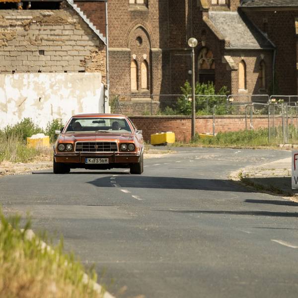 Ein roter Ford Gran Torino fährt mit Kölner Kennzeichen auf einer Straße entlang.