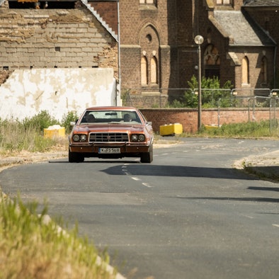Ein roter Ford Gran Torino fährt mit Kölner Kennzeichen auf einer Straße entlang.
