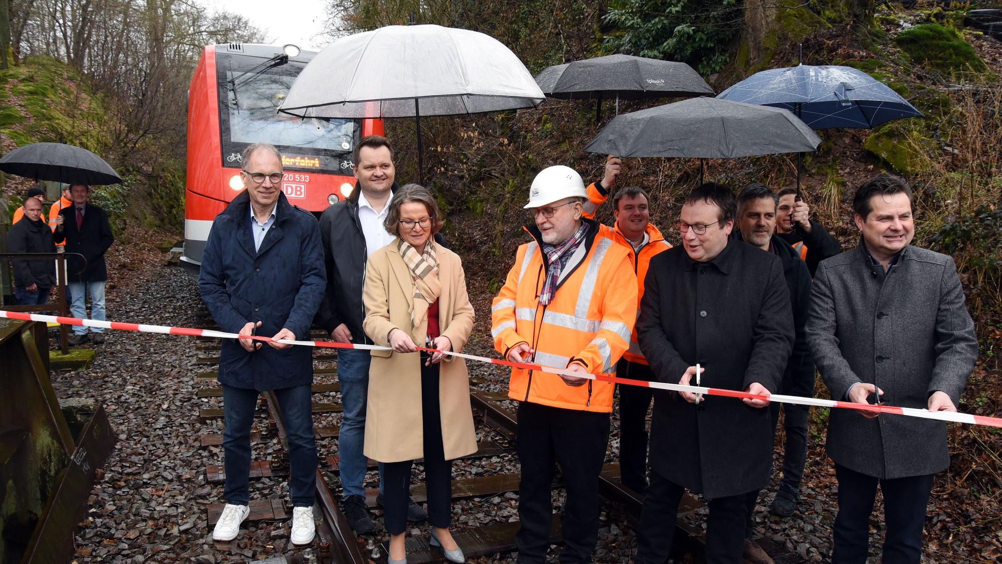 Eine Frau schneidet im Beisammensein vieler Männer ein Absperrband durch. Alle stehen auf Bahngleisen, im Hintergrund ist eine Regionalbahn zu sehen.