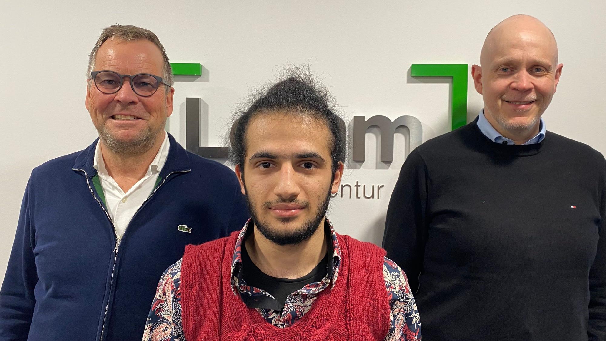 Mustapha steht mit den Geschäftsführern vor einer weißen Wand und blickt in die Kamera,