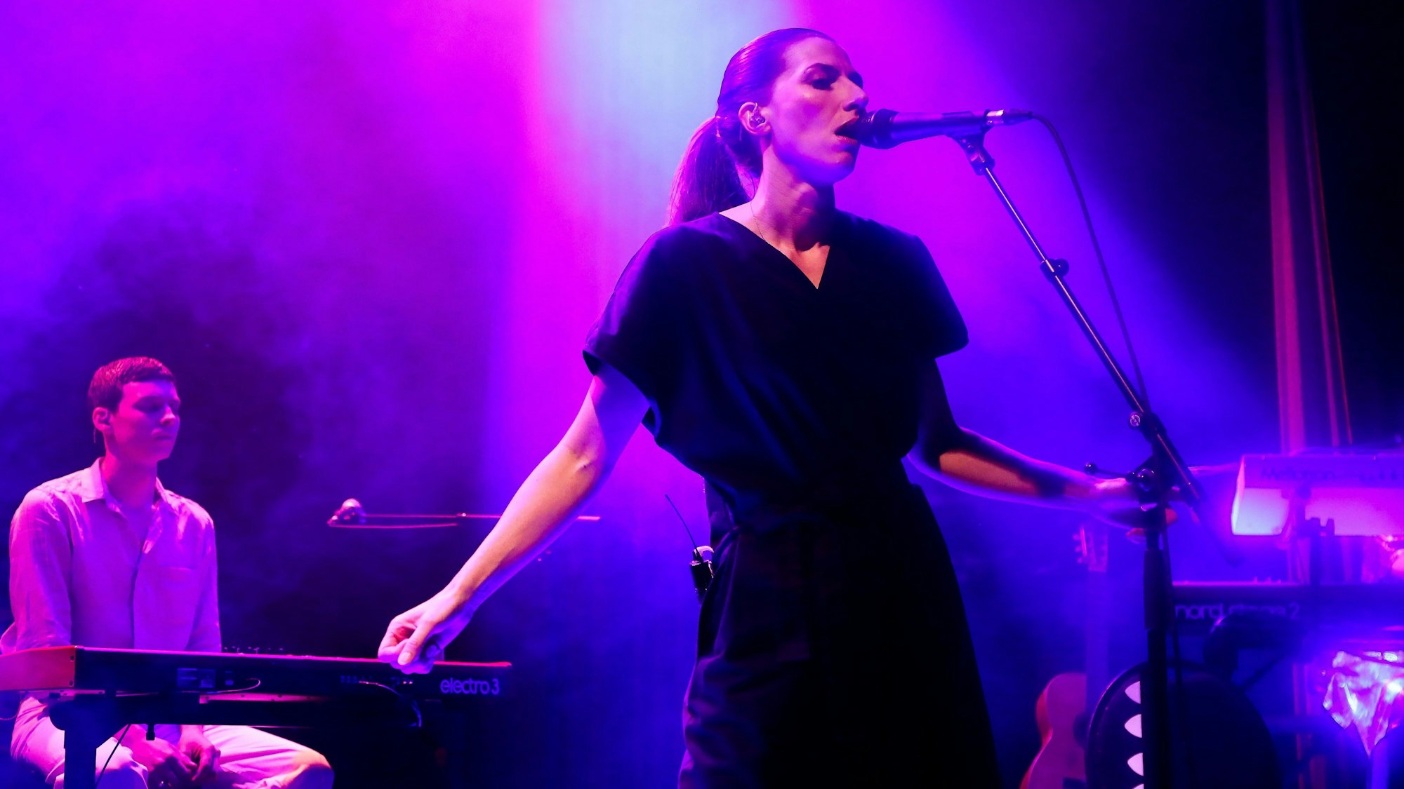 Aldous Harding singt im Kölner Gloria-Theater in Köln. Sie trägt einen ärmellosen schwarzen Hosenanzug und hat die Haare streng zum Zopf gebunden.
