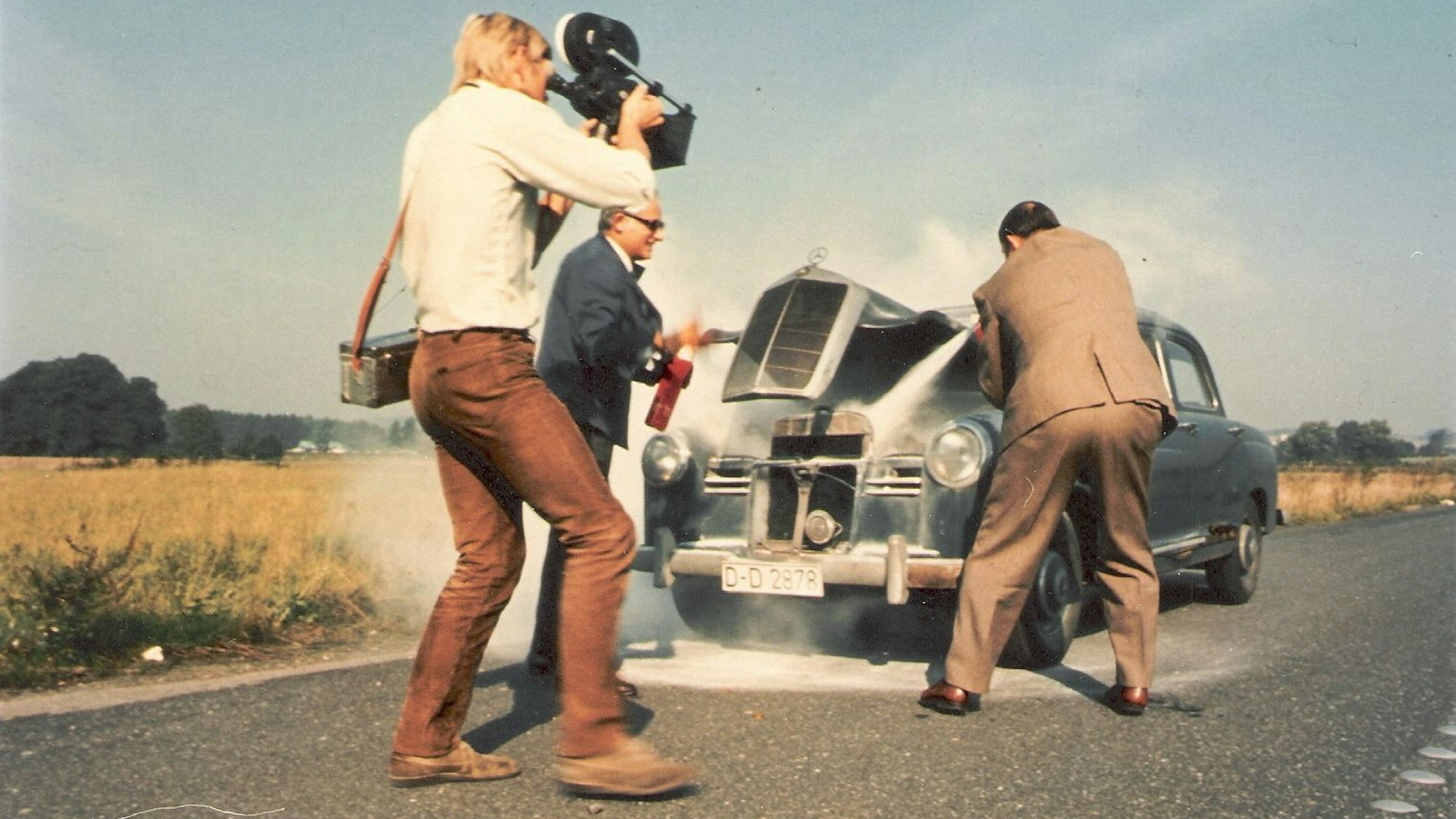 Zwei Männer löschen ein brennendes Auto, einer filmt.