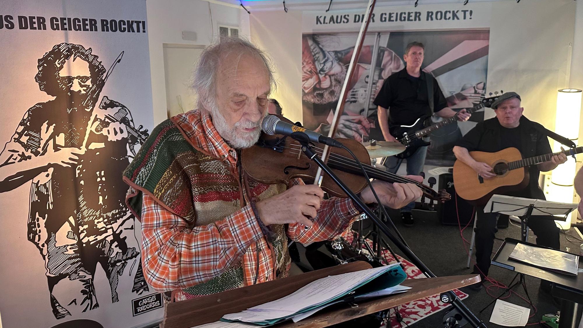 Mann, der mit Geige und Mikrofon vor einem Notenständer steht, im Hintergrund sieht man zwei weitere Musiker mit einer Gitarre und einer E-Gitarre.