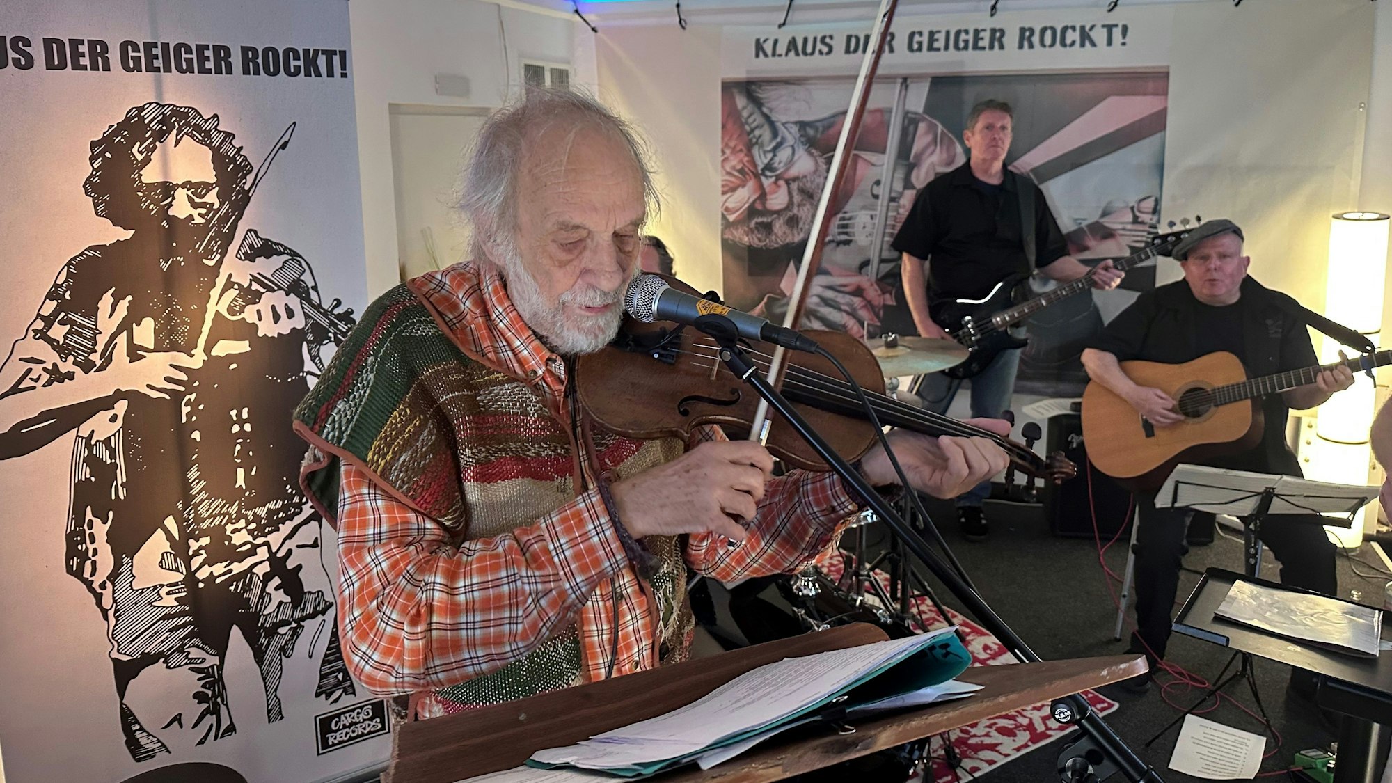Mann, der mit Geige und Mikrofon vor einem Notenständer steht, im Hintergrund sieht man zwei weitere Musiker mit einer Gitarre und einer E-Gitarre.