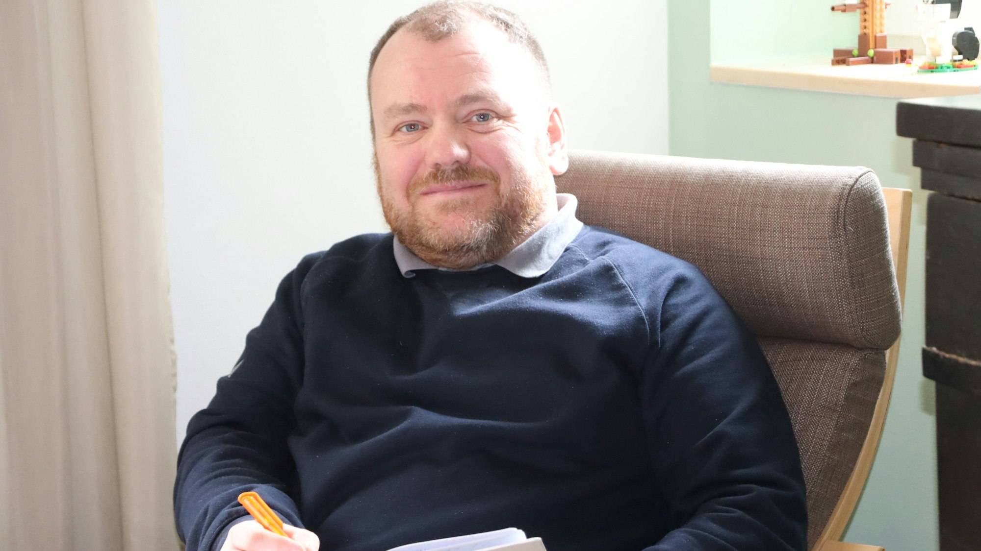 Ein Mann mit einem Notizbuch in der Hand sitzt auf einem Sessel.