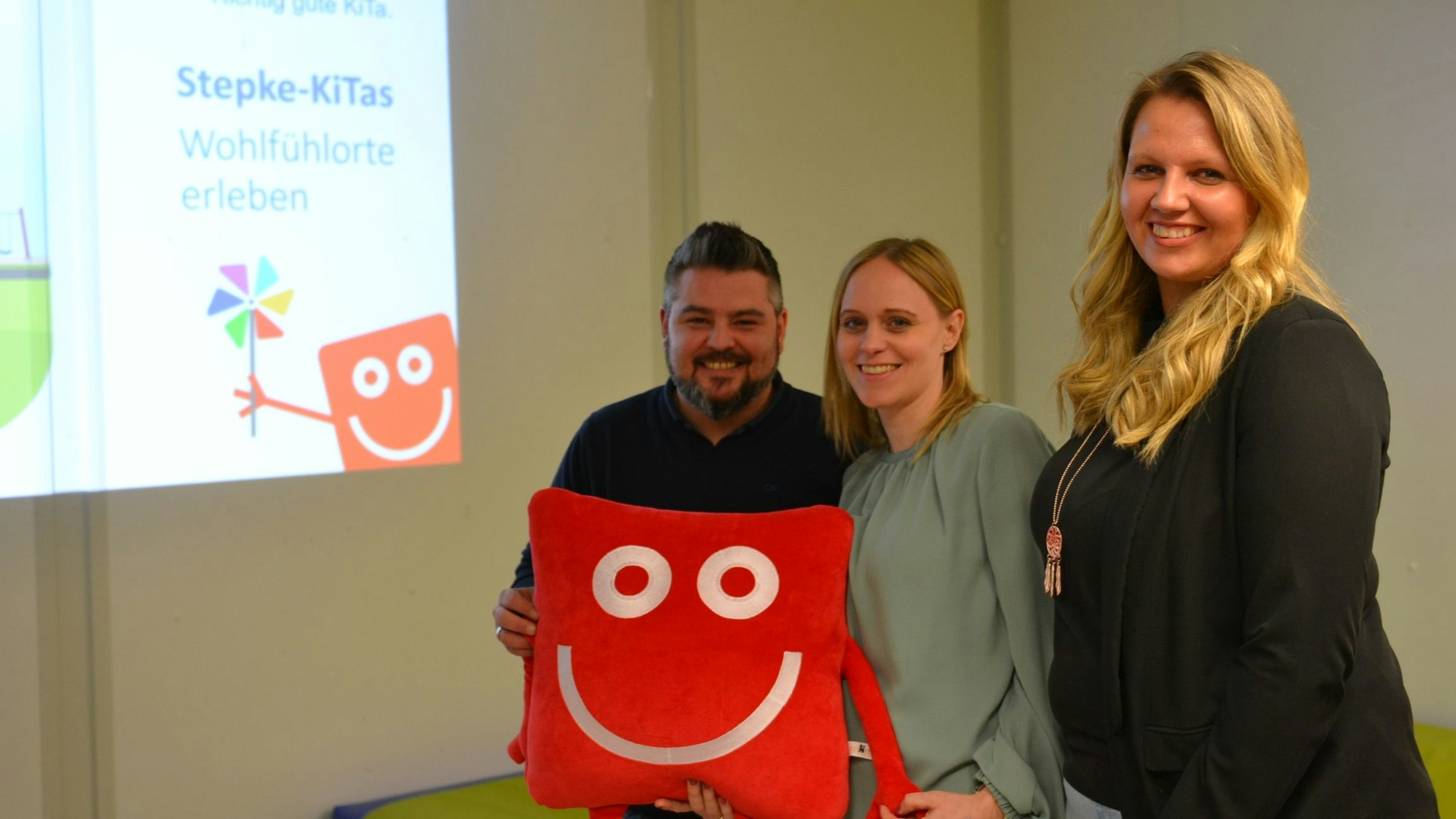 Die Leiter halten das rote, eckige Stoffmaskottchen in den Händen und lächeln in die Kamera.
