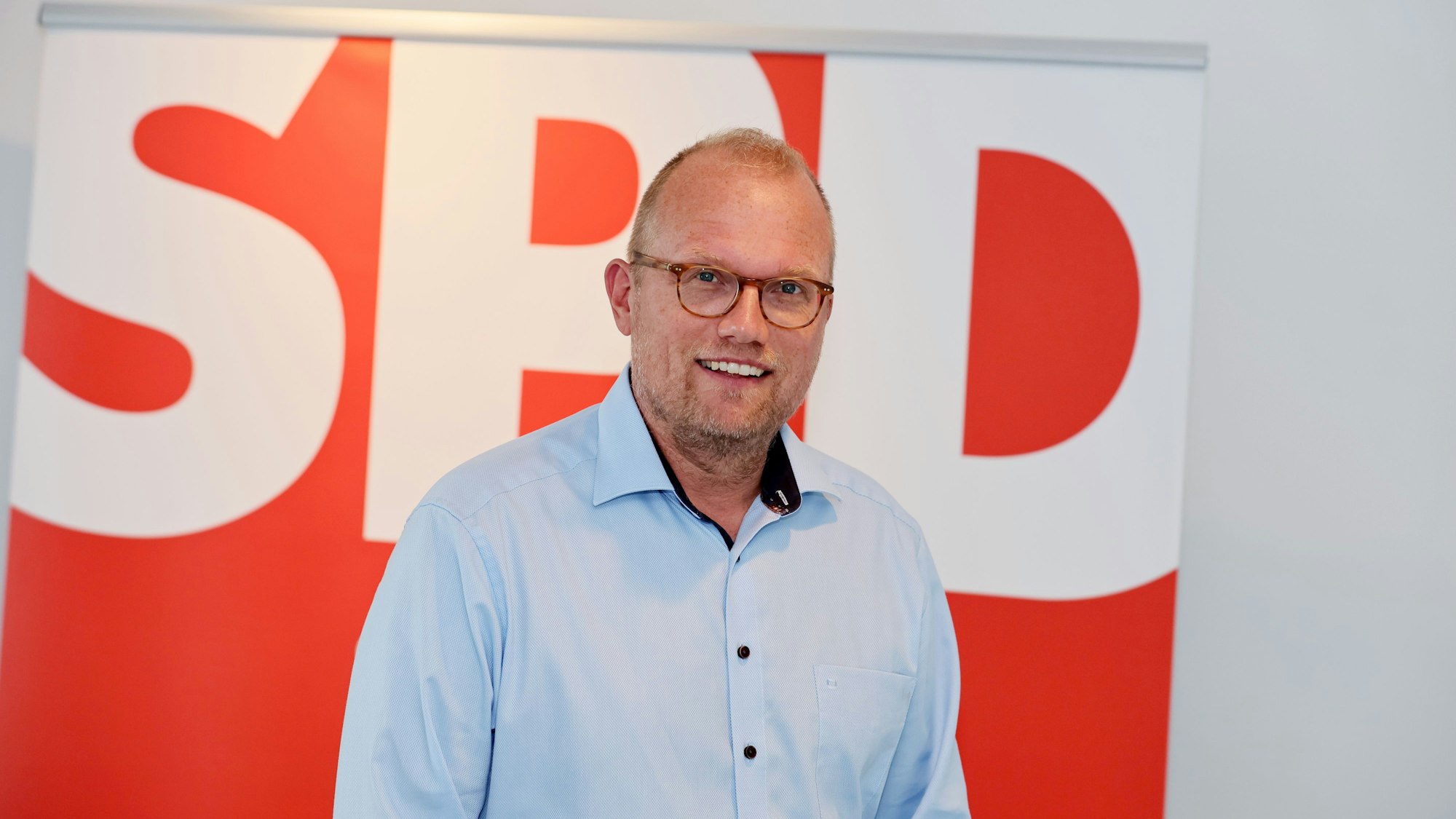 Jochen Ott steht vor einem SPD-Plakat und lächelt in die Kamera.