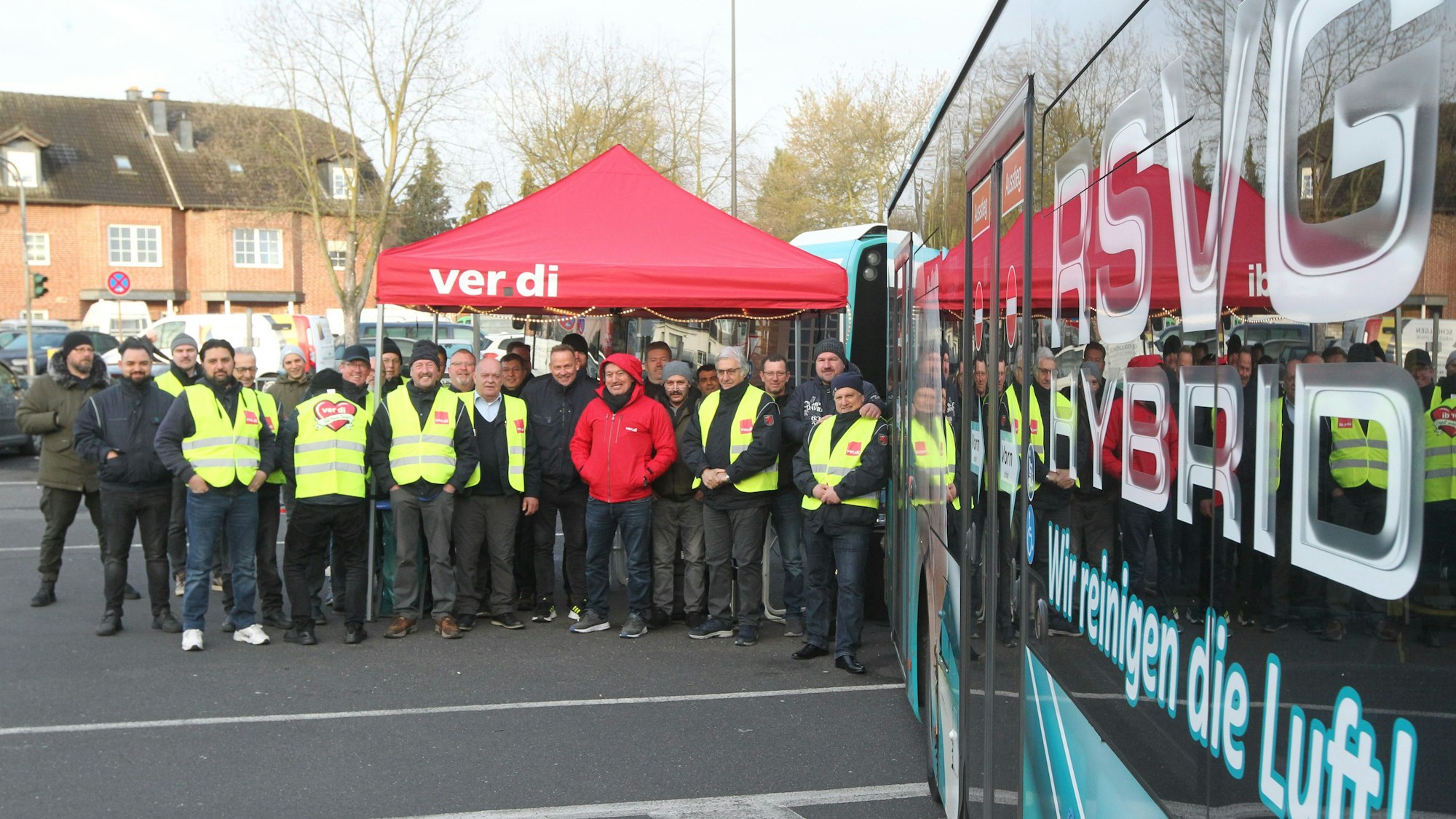 Männer in gelben Warnwesten der Gewerkschaft stehen unter einem Verdi-Pavillon neben einem Bus.
