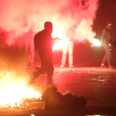 Gegenstände brennen auf einer Kreuzung, zwei vermummte Personen sind zu sehen.