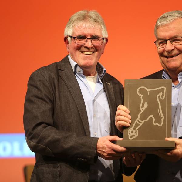 Zwei Männer stehen nebeneinander. In der Mitte halten sie gemeinsam einen eckigen Stein-Pokal in der Hand, in dessen Platte ein Fußballer eingraviert ist.