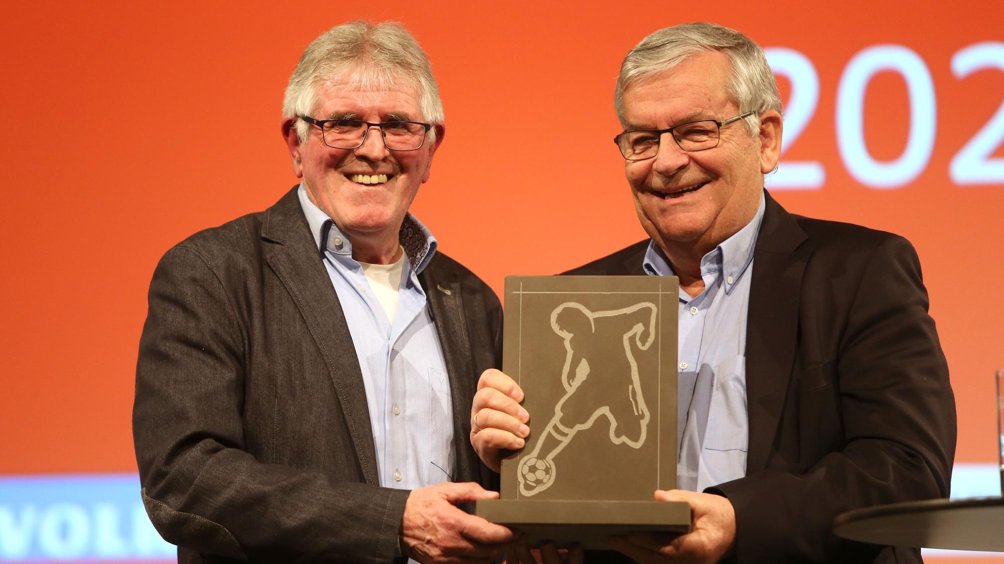 Zwei Männer stehen nebeneinander. In der Mitte halten sie gemeinsam einen eckigen Stein-Pokal in der Hand, in dessen Platte ein Fußballer eingraviert ist.