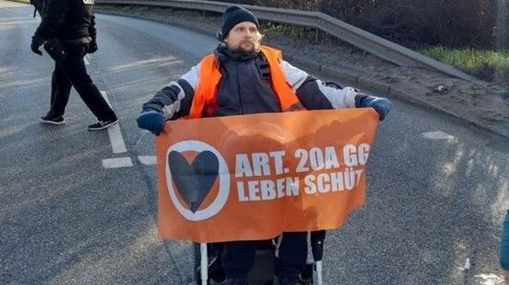 Ein Rollstuhlfahrer blockiert am Dienstag (28. März) eine Hauptverkehrsader am Hamburger Hafen. Er hält ein orangefarbenes Banner hoch.