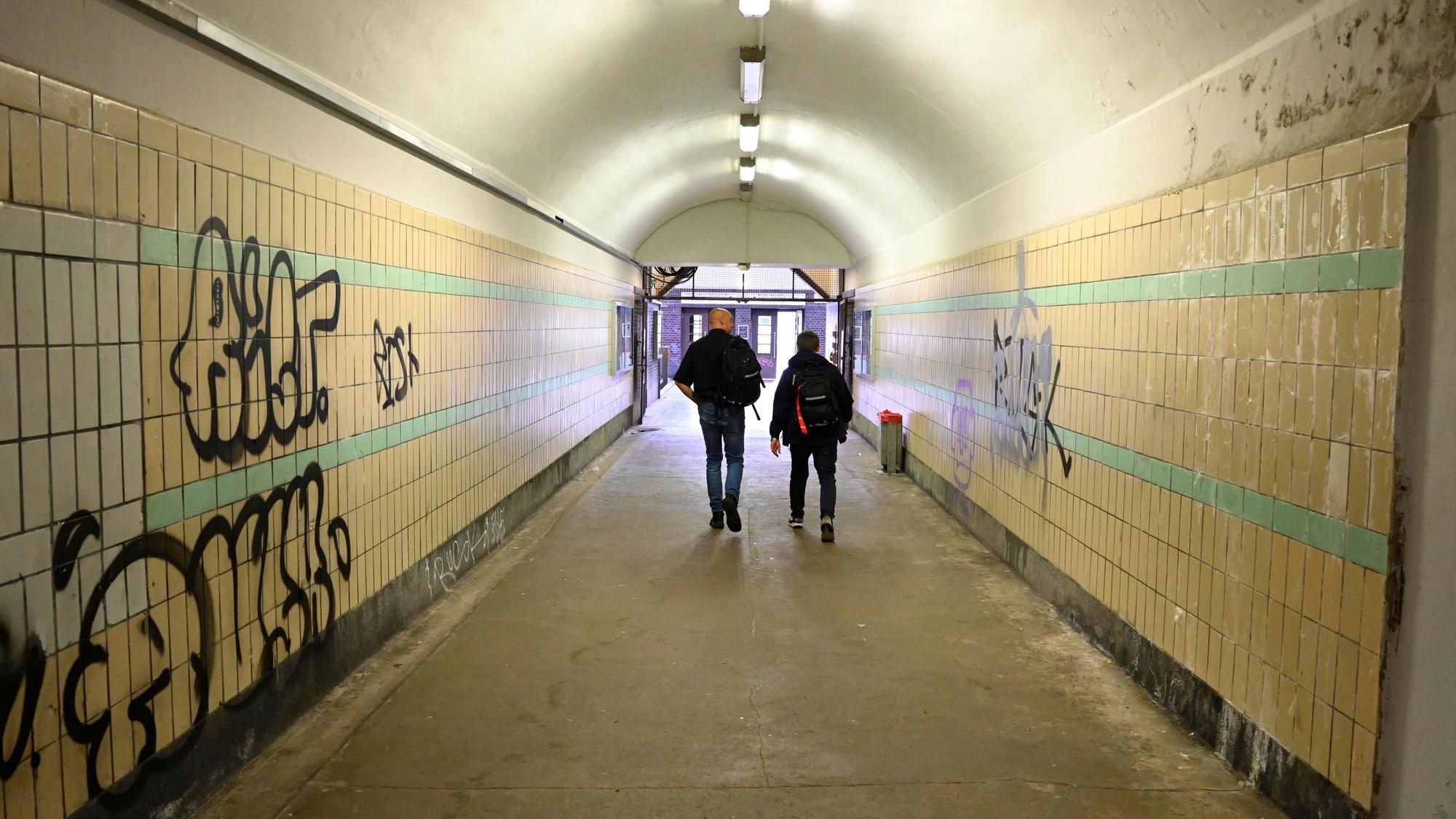 Zu sehen sind zwei Männer, die durch die Unterführung eines Bahnhofs gehen, deren Wände beschmiert sind.