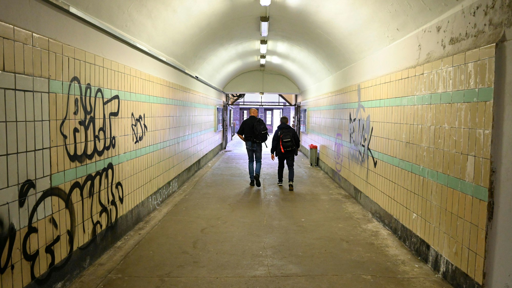 Zu sehen sind zwei Männer, die durch die Unterführung eines Bahnhofs gehen, deren Wände beschmiert sind.