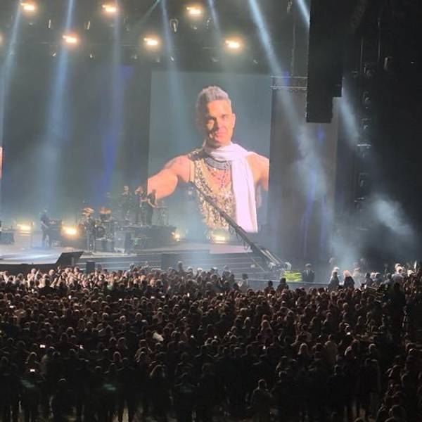 Eine Menschenmenge steht um eine Bühne. Auf großen LED-Wänden sieht man Robbie Williams.