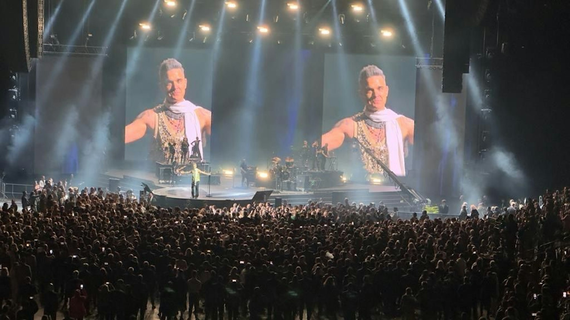 Eine Menschenmenge steht um eine Bühne. Auf großen LED-Wänden sieht man Robbie Williams.