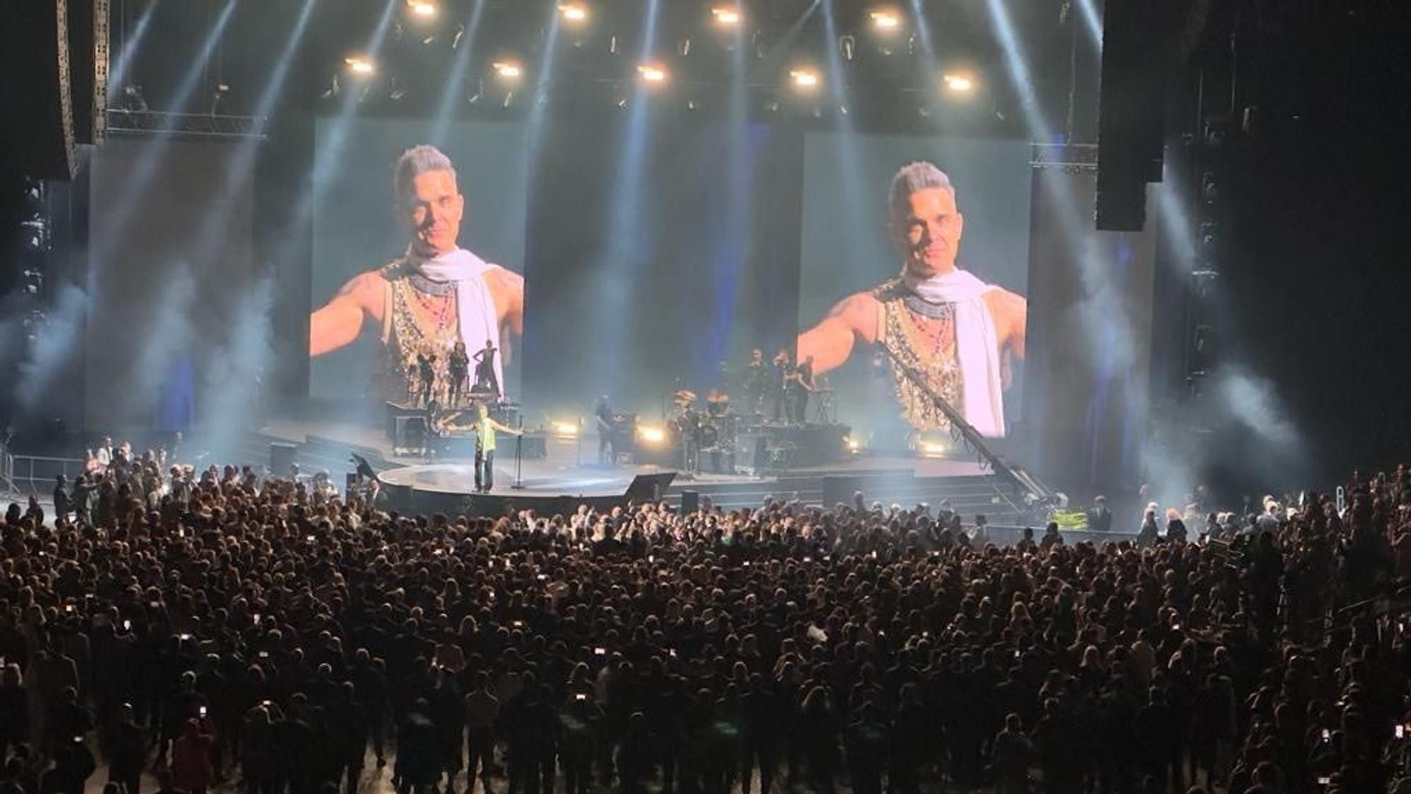 Eine Menschenmenge steht um eine Bühne. Auf großen LED-Wänden sieht man Robbie Williams.