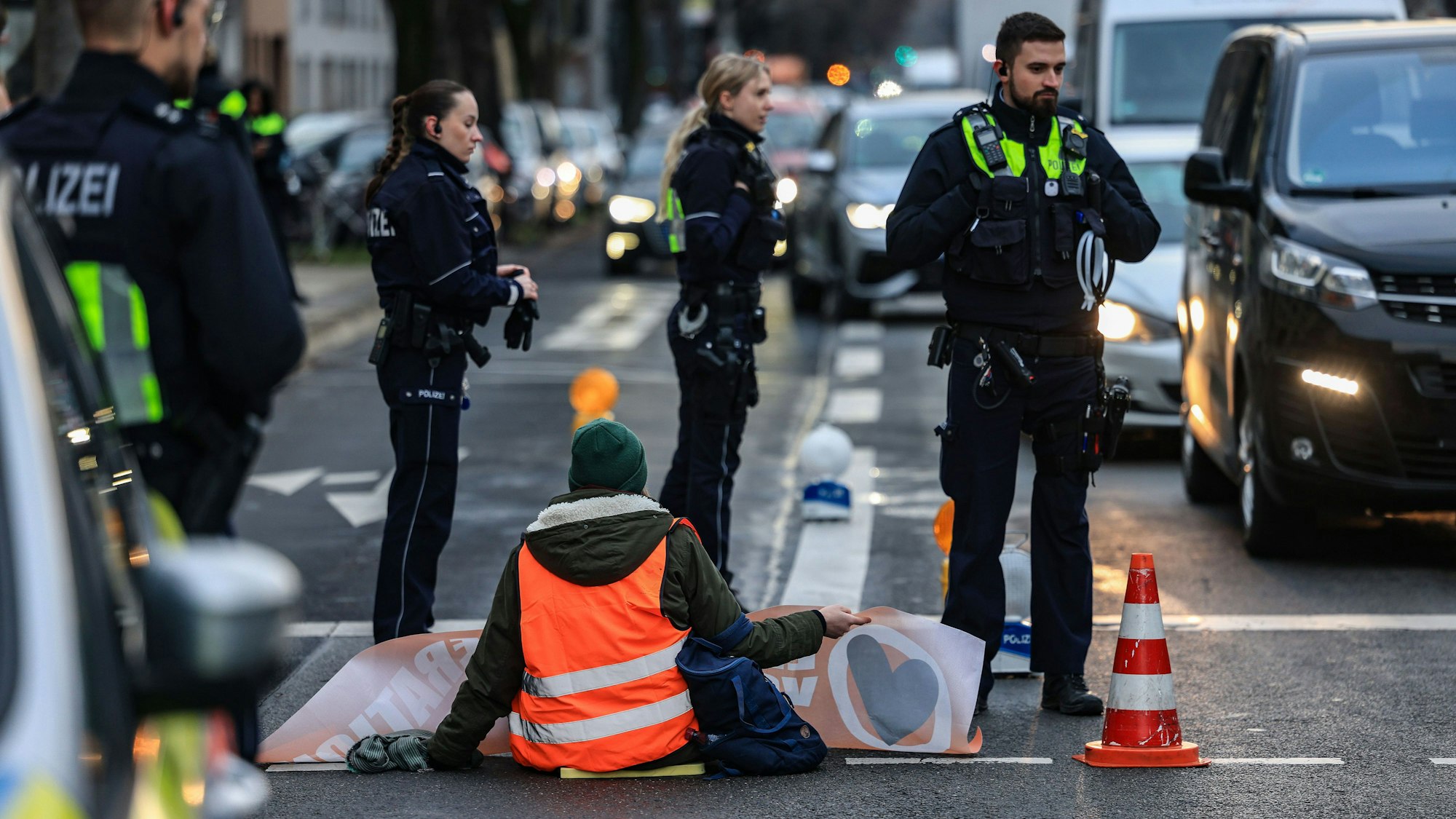 Auf der Fahrbahn sitzt eine Klimaaktivistin der „Letzten Generation“, mit dem Rücken zum Betrachter; eine Hand ist auf dem Untergrund festgeklebt, mit der anderen hält sie ein Plakat hoch. Vier junge Polizisten und Polizistinnen umringen sie, um sie vor dem Autoverkehr zu schützen.