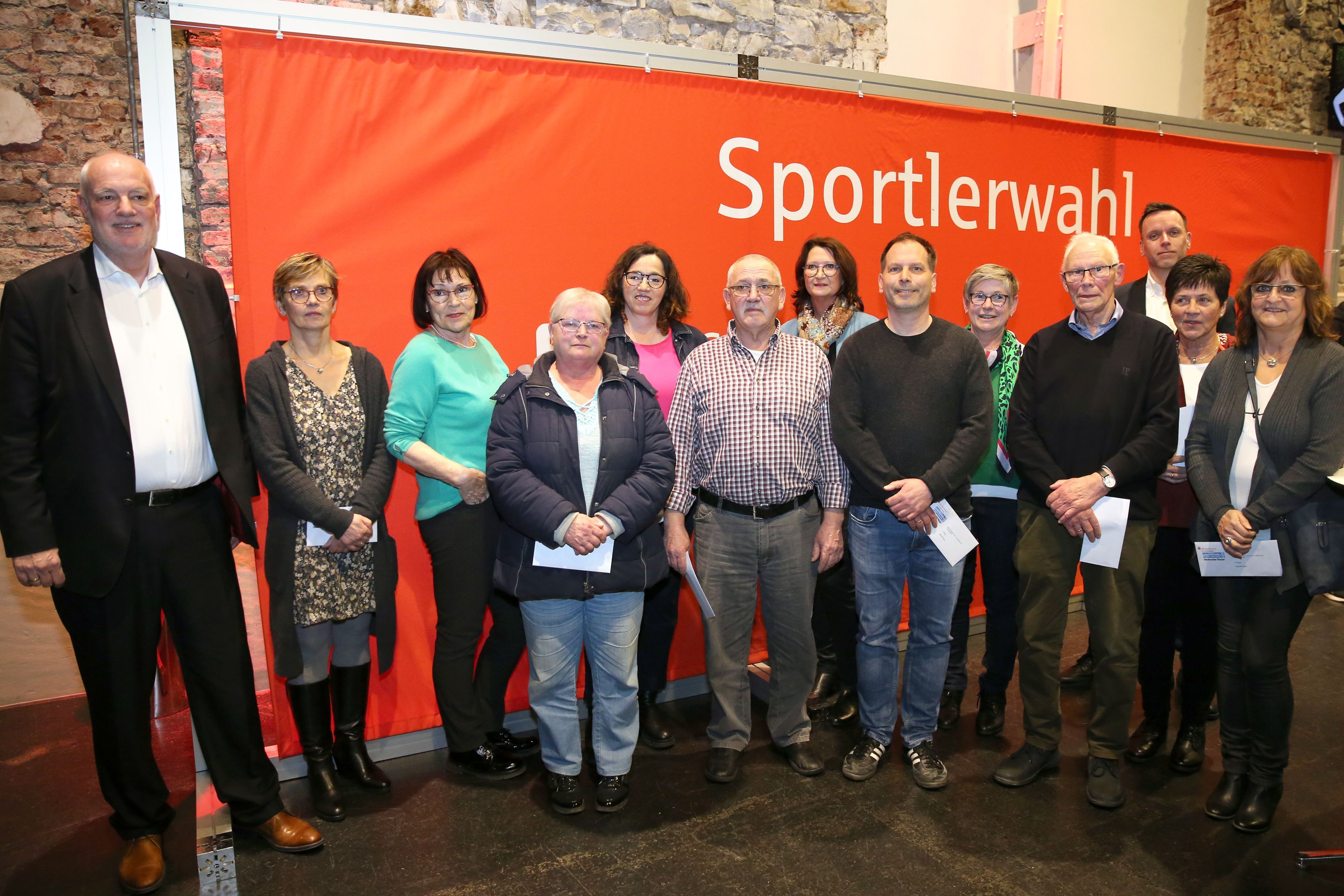 Auf einem Gruppenfoto stehen mehrere Frauen und Männer gemeinsam vor einem roten Banner mit der weißen Aufschrift „Sportlerwahl“.