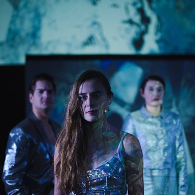 Ein Schauspieler und zwei Schauspielerinnen stehen in blau glänzenden Kostümen auf der Bühne.