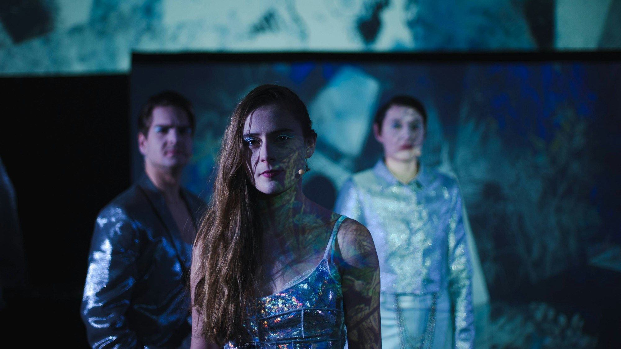 Ein Schauspieler und zwei Schauspielerinnen stehen in blau glänzenden Kostümen auf der Bühne.