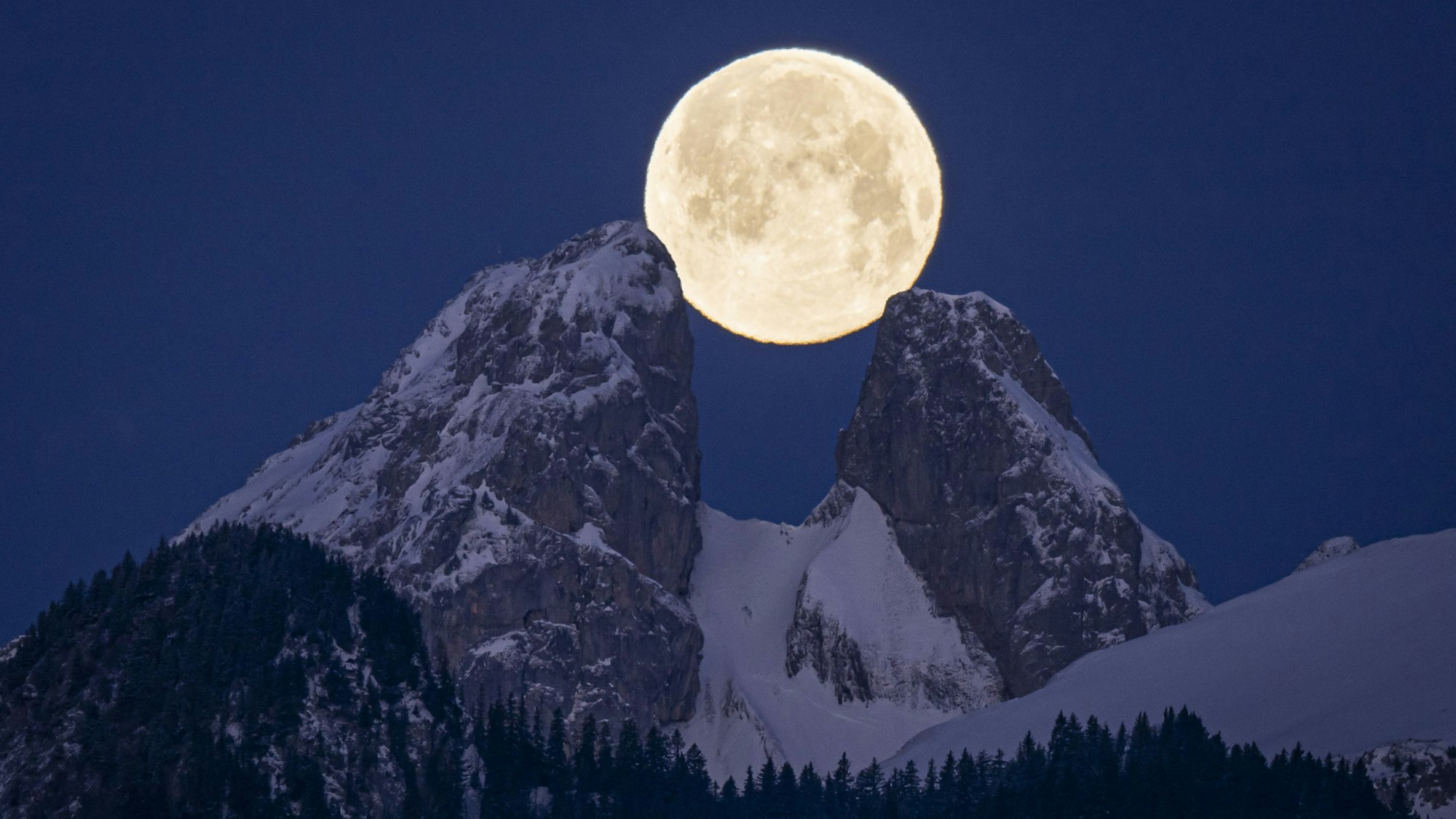 Der Vollmond geht hinter zwei verschneiten Berggipfeln auf. Er ist deutlich am klaren und tiefblauen Himmel zu sehen.