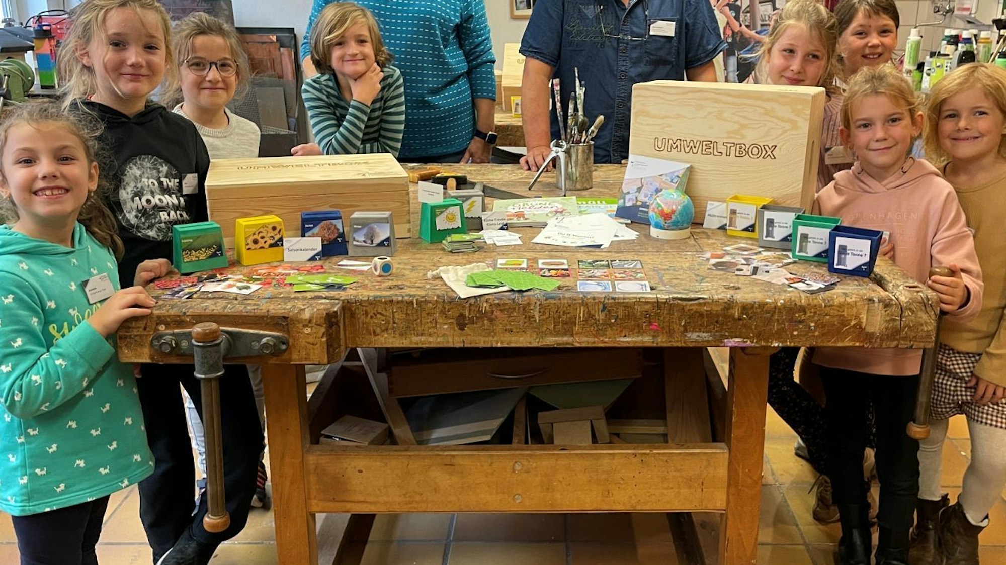 Acht Kinder stehen um einen Holztisch, auf dem die Inhalte der Umweltbox verteilt sind, Karten-, Malspiele, Infoblätter und vieles mehr.