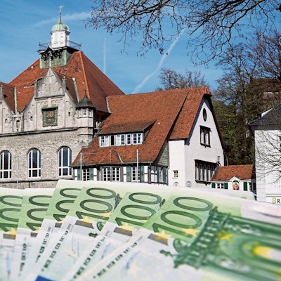 Hundert-Euro-Scheine aufgefächert im Vordergrund, im Hintergrund das Rathaus in Bergisch Gladbach.