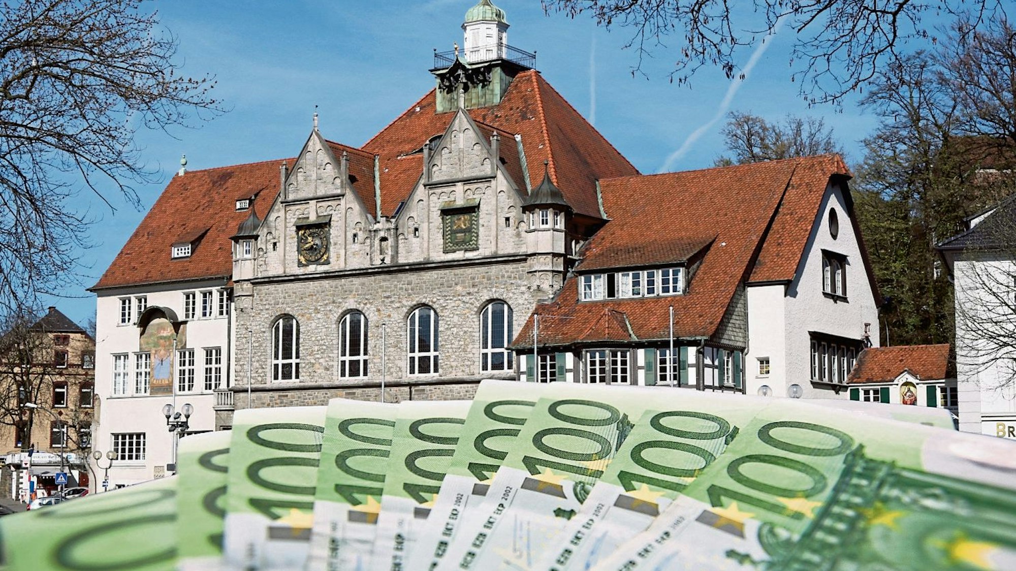 Hundert-Euro-Scheine aufgefächert im Vordergrund, im Hintergrund das Rathaus in Bergisch Gladbach.