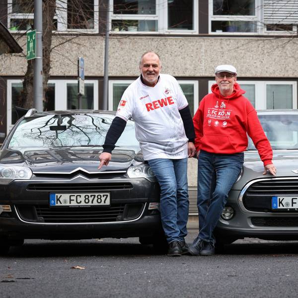 Zwei Männer in Fußballtrikots stehen vor ihren Autos und zeigen auf das Nummernschild.