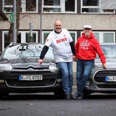 Zwei Männer in Fußballtrikots stehen vor ihren Autos und zeigen auf das Nummernschild.