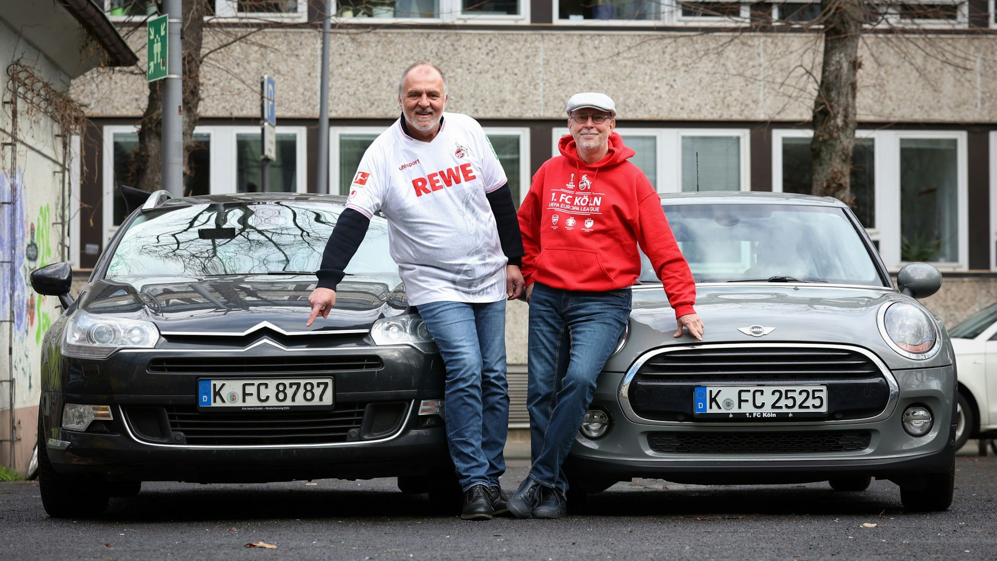 Zwei Männer in Fußballtrikots stehen vor ihren Autos und zeigen auf das Nummernschild.