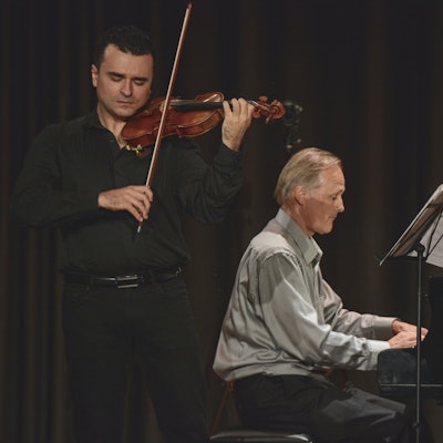 Zwei Personen bei einem Auftritt, ein Mann spielt Violine und ein anderer Mann Klavier.