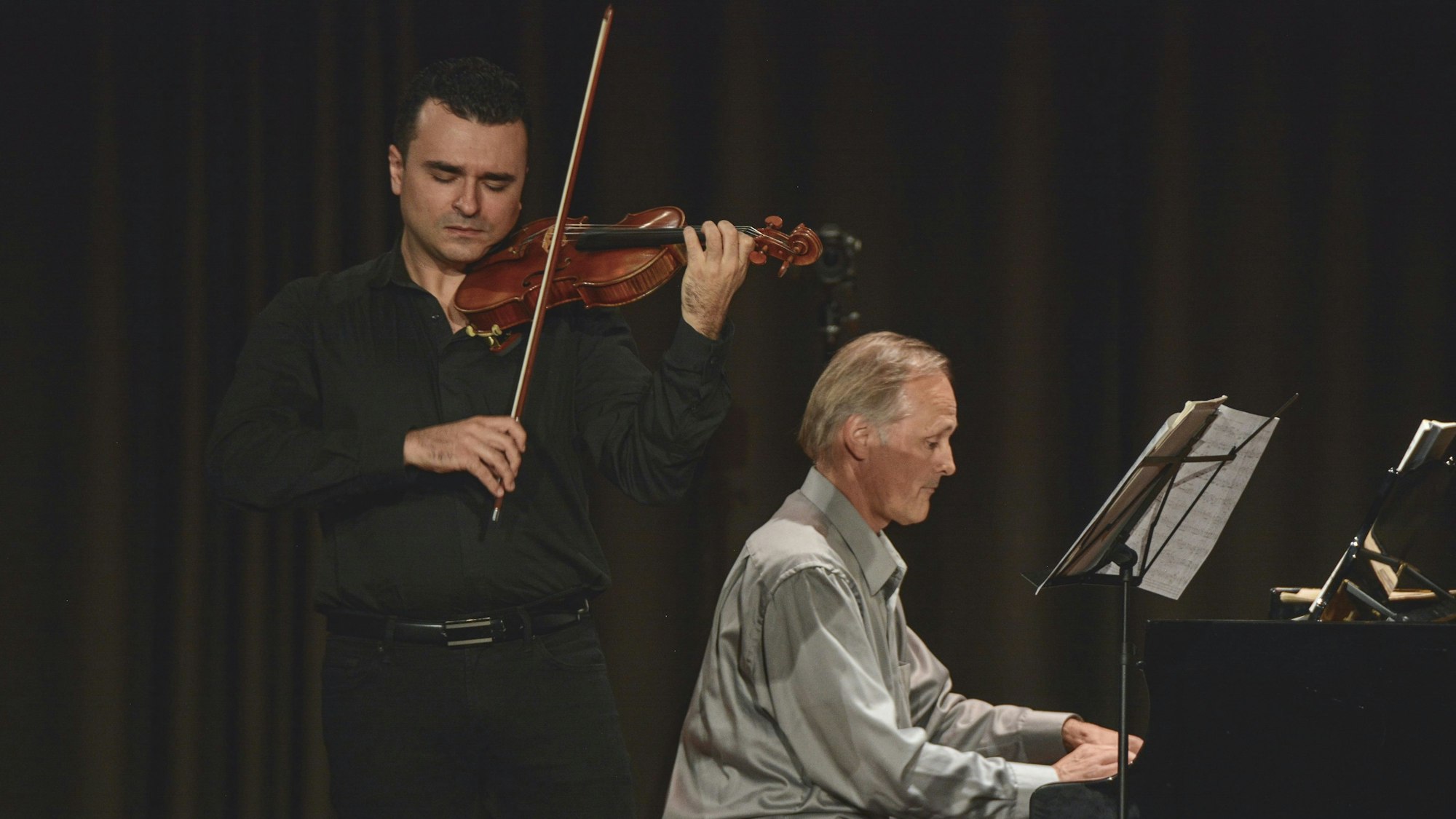 Zwei Personen bei einem Auftritt, ein Mann spielt Violine und ein anderer Mann Klavier.