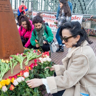 Erinnerung an den Genozid an Armeniern am Heinrich-Böll-Platz: Eine Frau mit Sonnenbrille legt Blumen vor einer Gedenkstele ab. Im Hintergrund ist die Hohenzollernbrücke zu sehen.