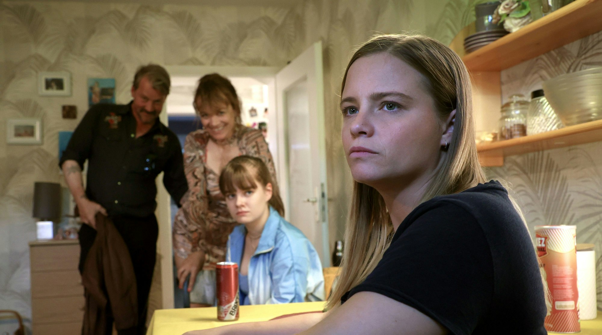 Szene aus „Donuts“, dem neuen „Tatort“ aus Bremen. Liv Moormann (Jasna Fritzi Bauer) sitzt am Tisch mit ihrer Schwester Marie (Luise Böse) und schaut nachdenklich und verzweifelt an der Kamera vor bei. Sie sitzt im Vordergrund. Hinter ihr sind verschwommen ihre Mutter Jenny (Angelika Richter), Marie und ein Mann zu sehen.