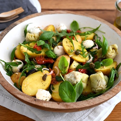 In einer Schüssel sind Kartoffeln, Rucola, Mozzarella, Artischocken, Oliven und getrocknete Tomaten zu einem Salat zubereitet.