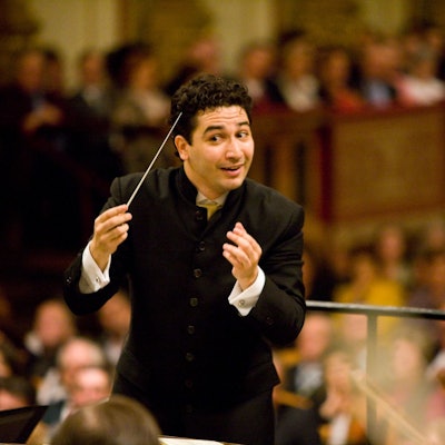 Andrés Orozco-Estradadirigitert mit einem Lächeln und hat einen Taktstock in der rechten Hand. Er trägt schwarze Kleidung.