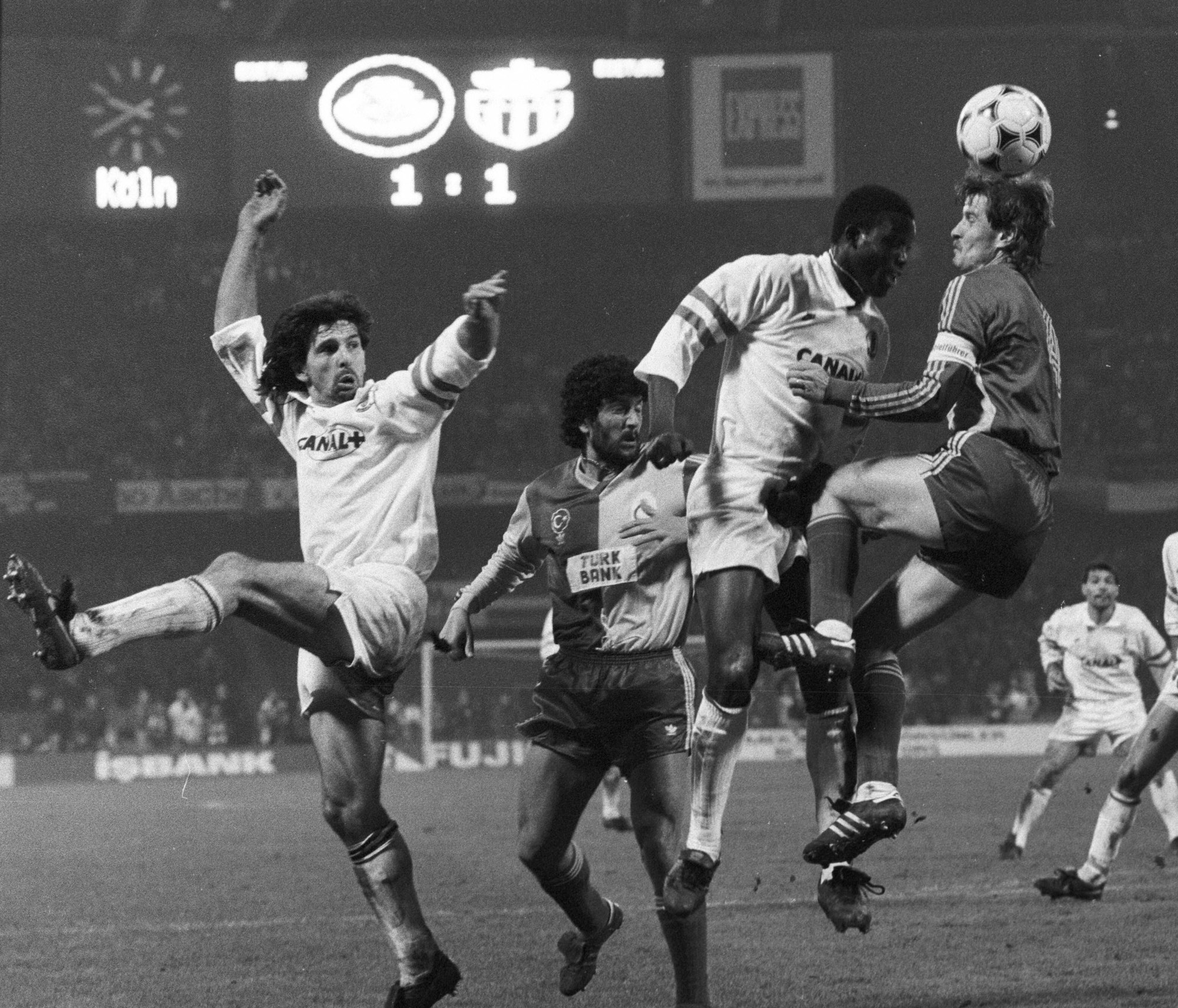 Auf dem Schwarz-Weiß-Bild kämpfen mehrere Fußballer beim Kopfball um das Spielgerät. Im Hintergrund leuchtet die große Anzeigentafel, auf der unter anderem das Wort "Köln" zu lesen ist.