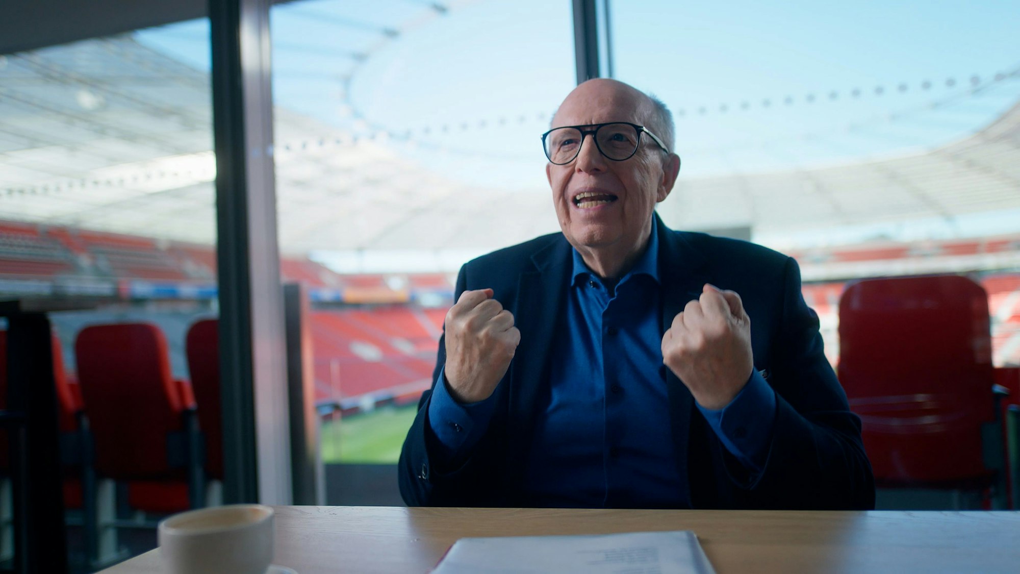 Reiner Calmund sitzt an einem Schreibtisch in einer Loge der BayArena Leverkusen. Teile des Stadions sind hinter ihm zu erkennen, er trägt eine schwarze Brille, ein dunkles Sakko sowie ein blaues Hemd.