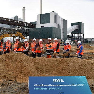 Mehrere Bauarbeiter stehen hinter einer Grube. Im Hintergrund das Goldenbergkraftwerk.