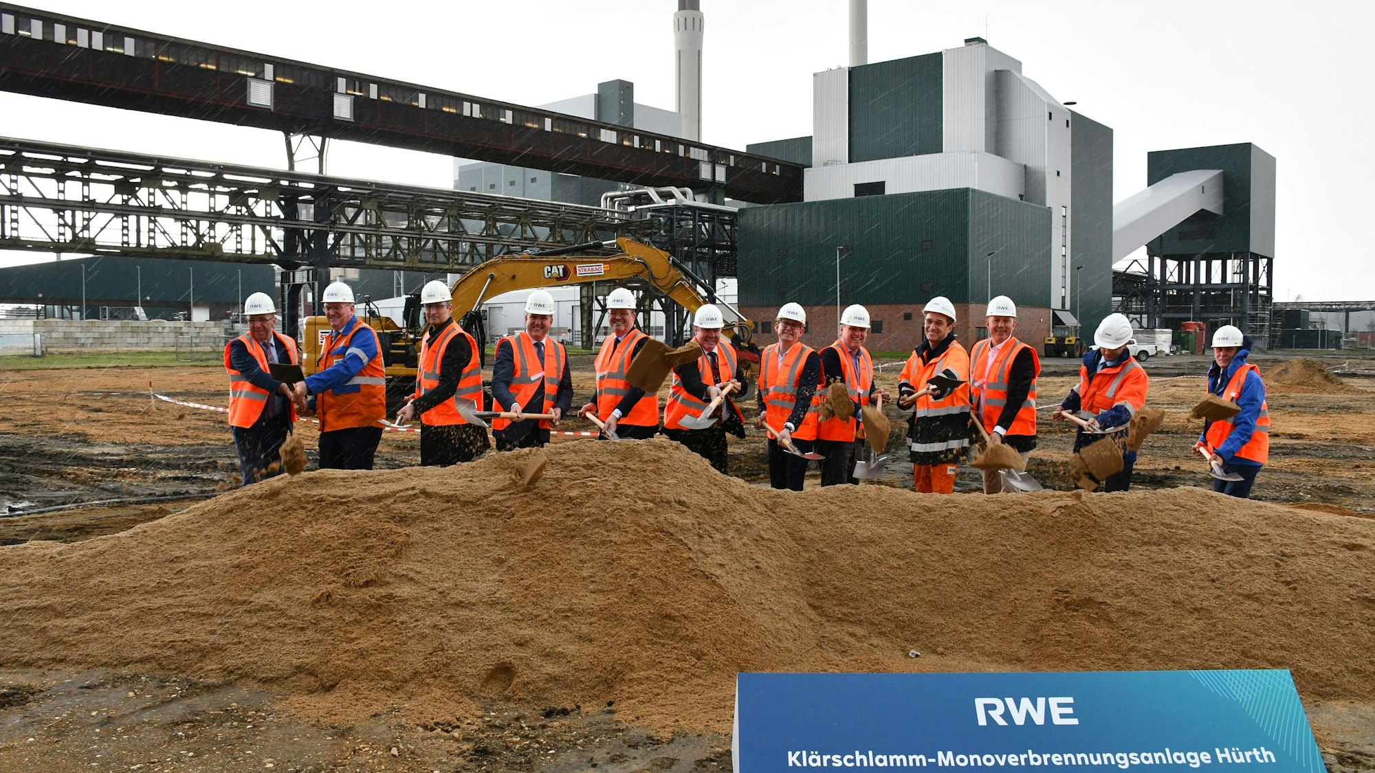 Mehrere Bauarbeiter stehen hinter einer Grube. Im Hintergrund das Goldenbergkraftwerk.