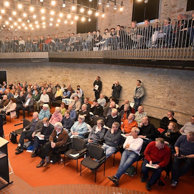 Menschen sitzen im Bensberger Ratssaal und hören einem Sprecher auf der Bühne zu.