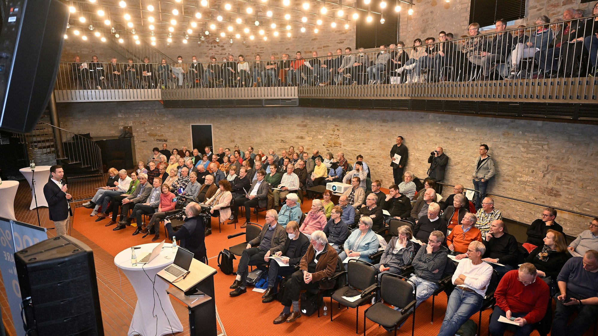 Menschen sitzen im Bensberger Ratssaal und hören einem Sprecher auf der Bühne zu.