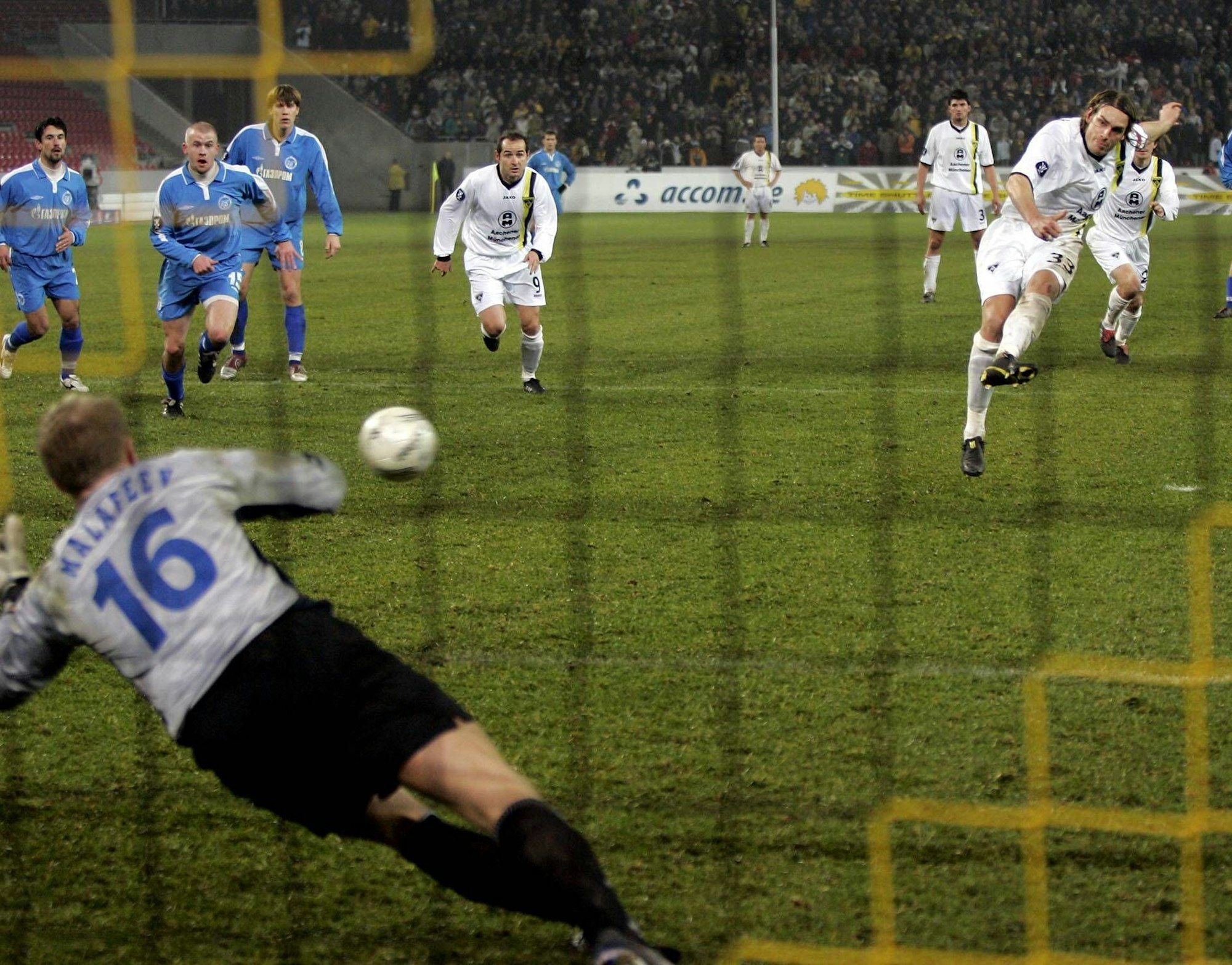 Ein ganz in weiß gekleideter Feldspieler tritt den Ball Richtung Tor. Der Torwart im Bildervordergrund versucht den Ball zu erreichen.