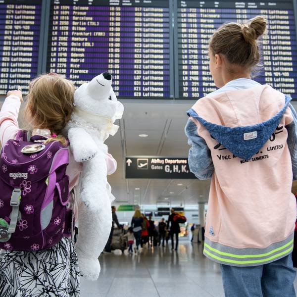 Zwei Mädchen blicken auf eine Abflugtafel am Flughafen. Eines trägt einen weißen Teddy im Arm.